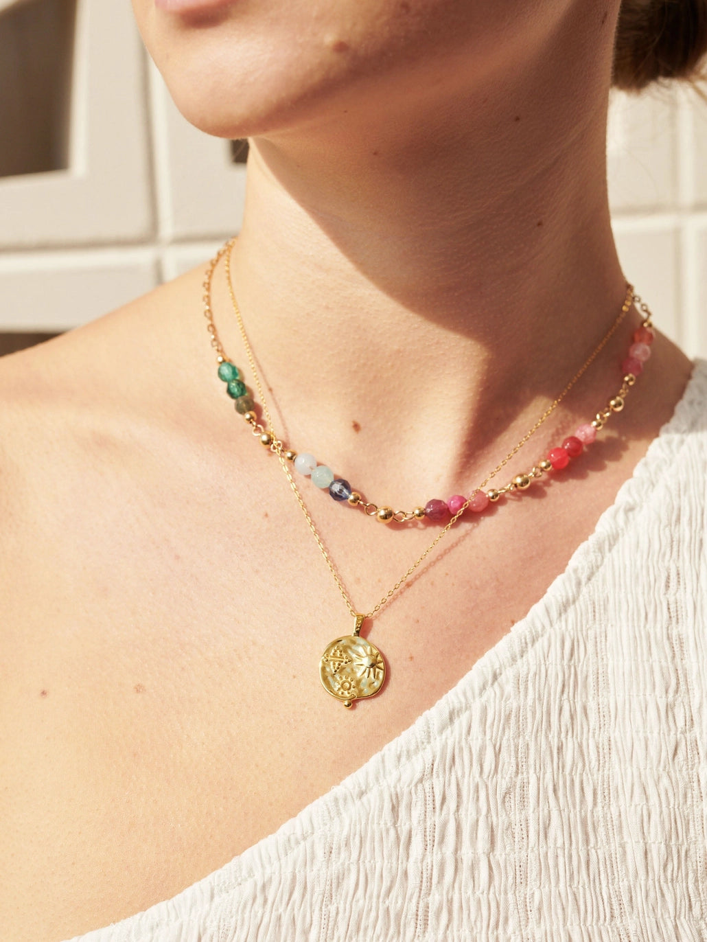 Layered gold medallion necklace with embossed sun and floral symbols styled with a colourful beaded choker, shown on a model’s neckline in natural sunlight.