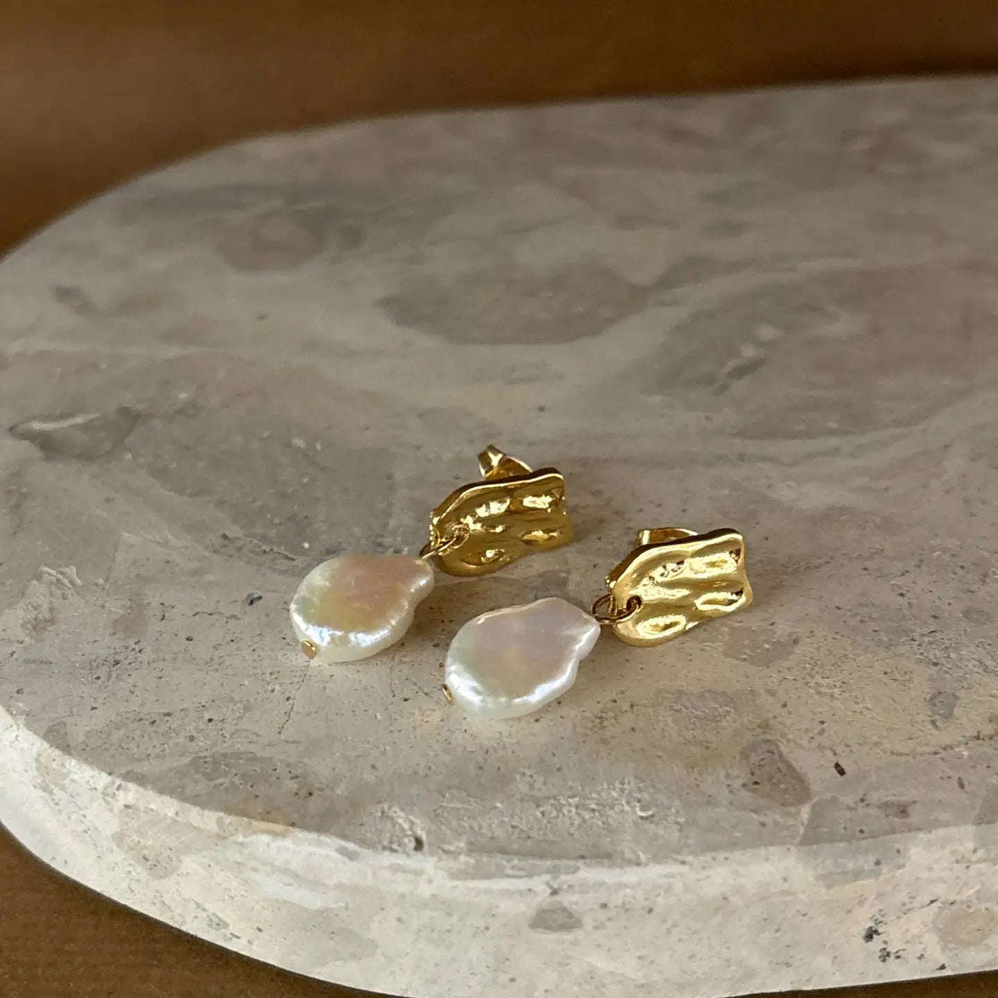 Close-up of gold stud earrings with baroque-style pearls resting on a neutral stone background.