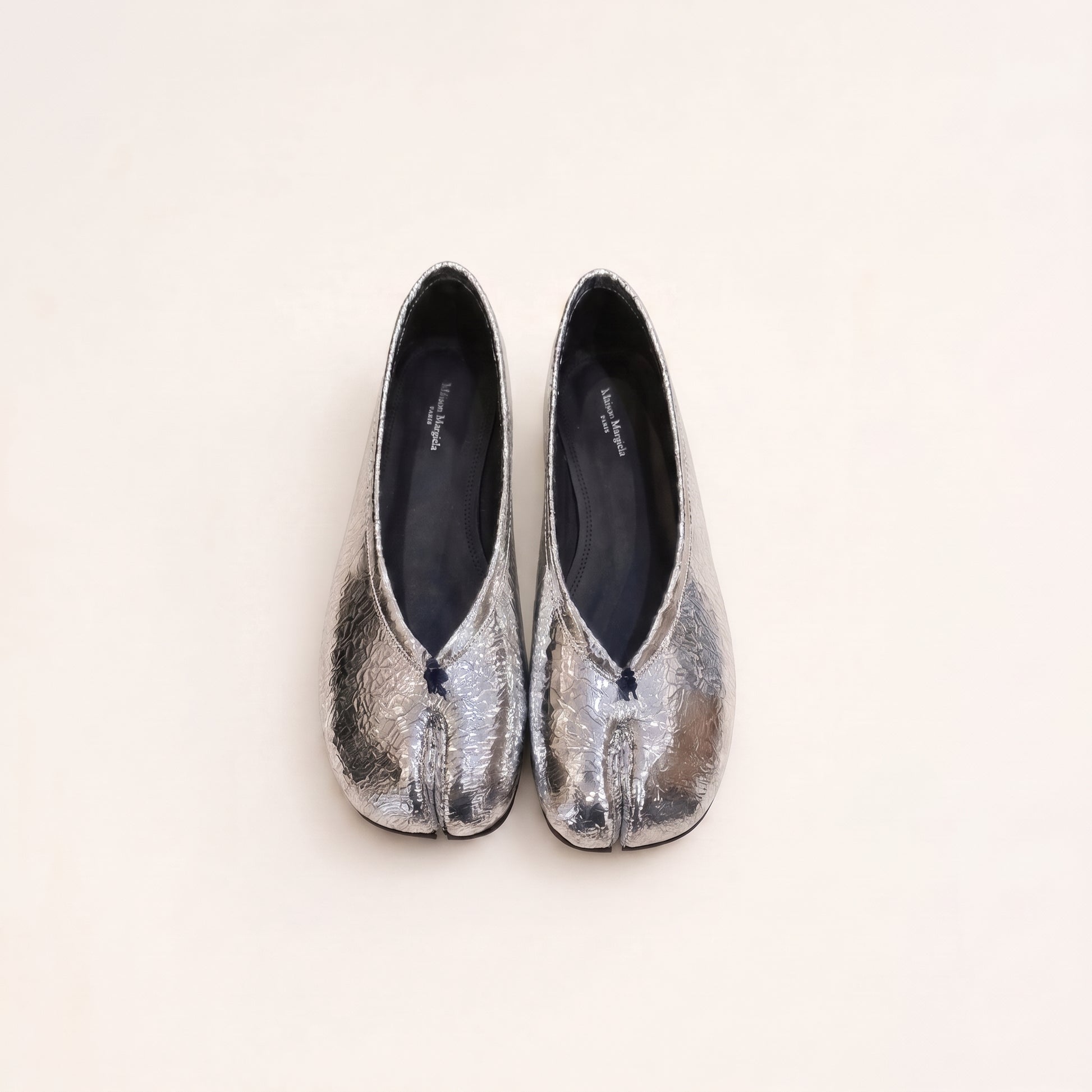 A pair of metallic silver tabi-style shoes with a split toe and small dark tassel details, photographed from above on a plain off-white background.