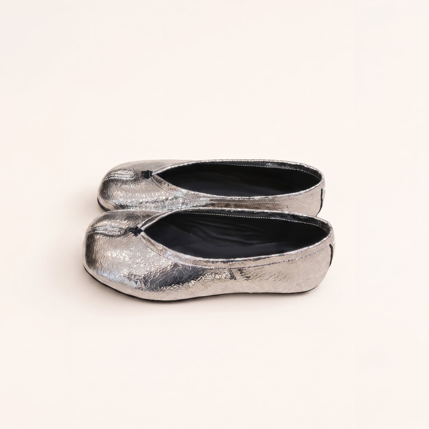 A pair of metallic silver flat shoes with a textured, crinkled finish and black interiors, photographed on a clean, solid off-white background.