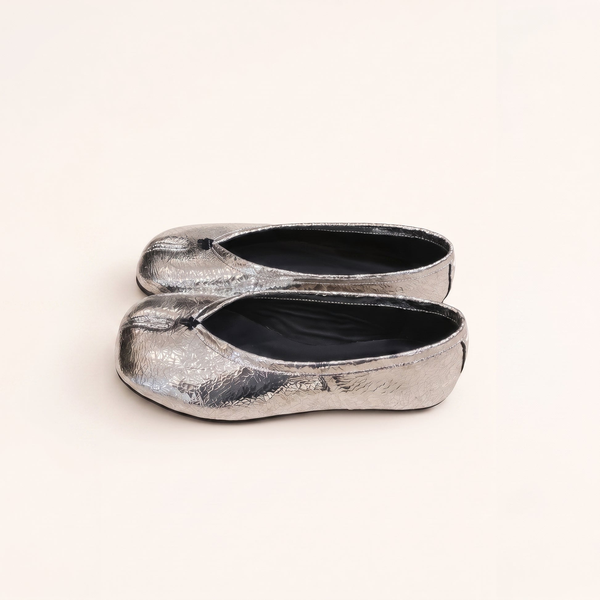 A pair of metallic silver flat shoes with a textured, crinkled finish and black interiors, photographed on a clean, solid off-white background.