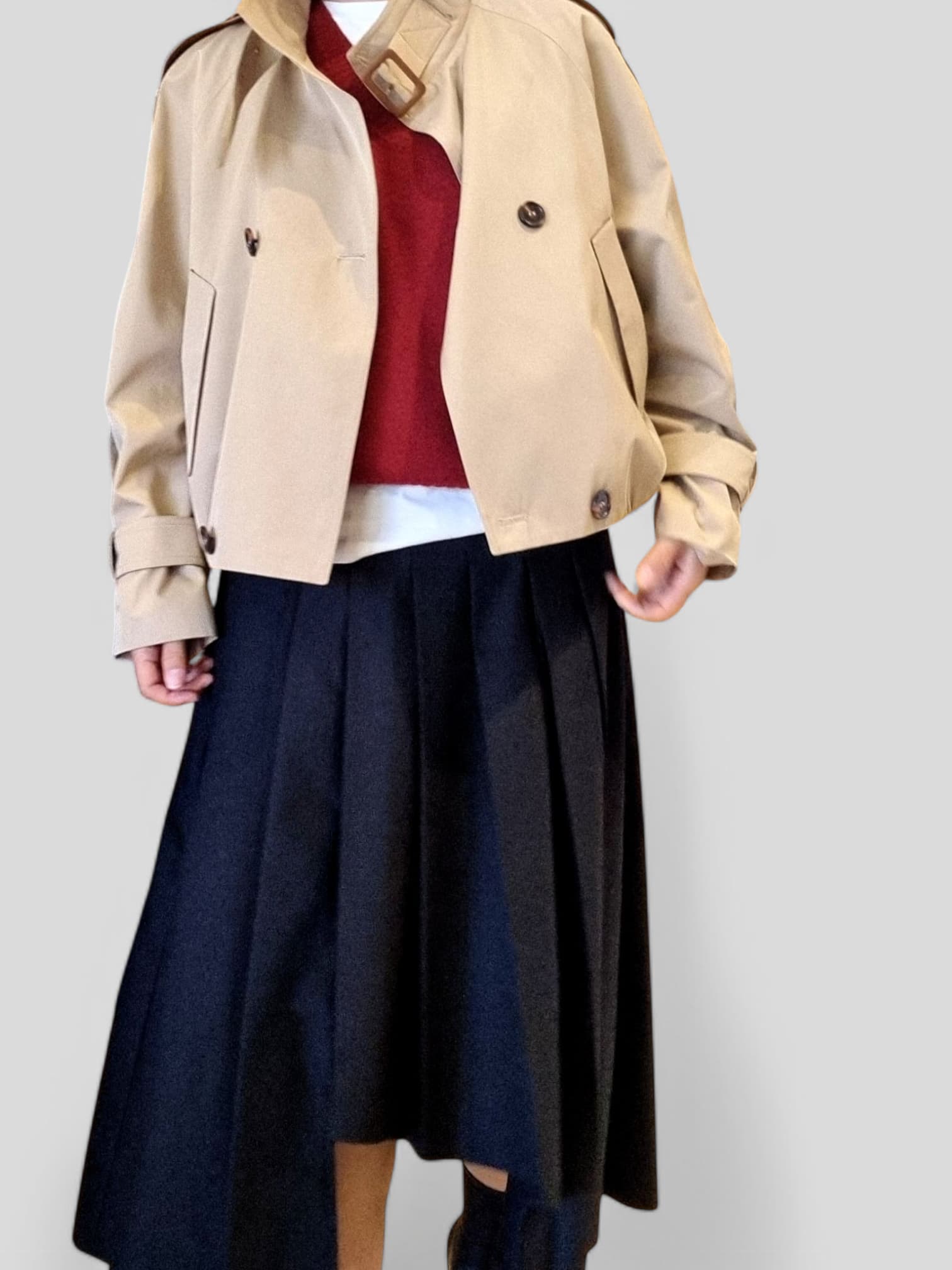 “Beige cropped trench coat styled with a pleated navy midi skirt and layered knit top on a neutral background.”