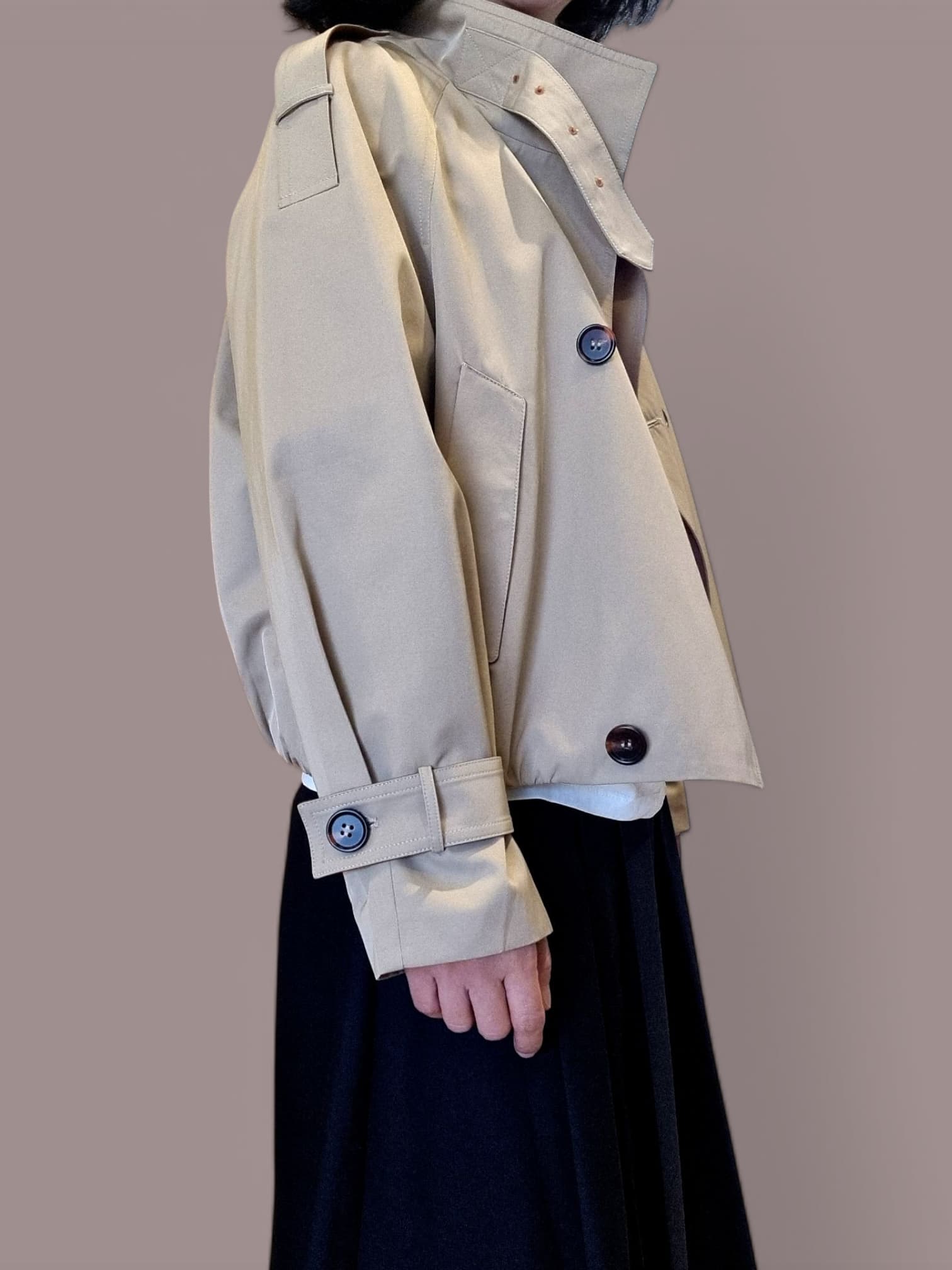 “Side view of a beige cropped trench coat with button details and cuff straps, styled over a dark pleated skirt on a neutral background.”