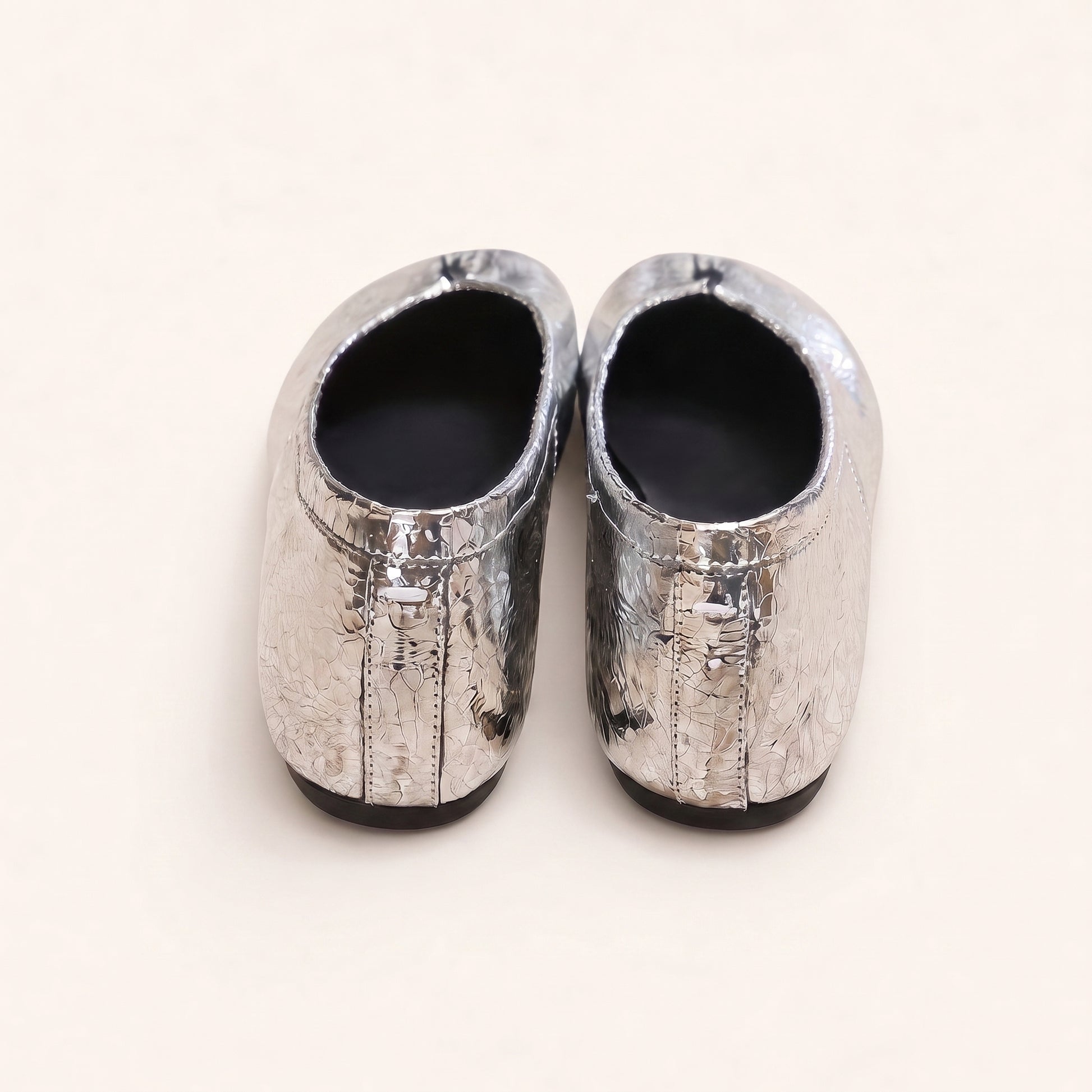 A pair of metallic silver slip-on shoes with a textured, cracked leather-like finish and black soles, shown from a top-down view on a light cream background.
