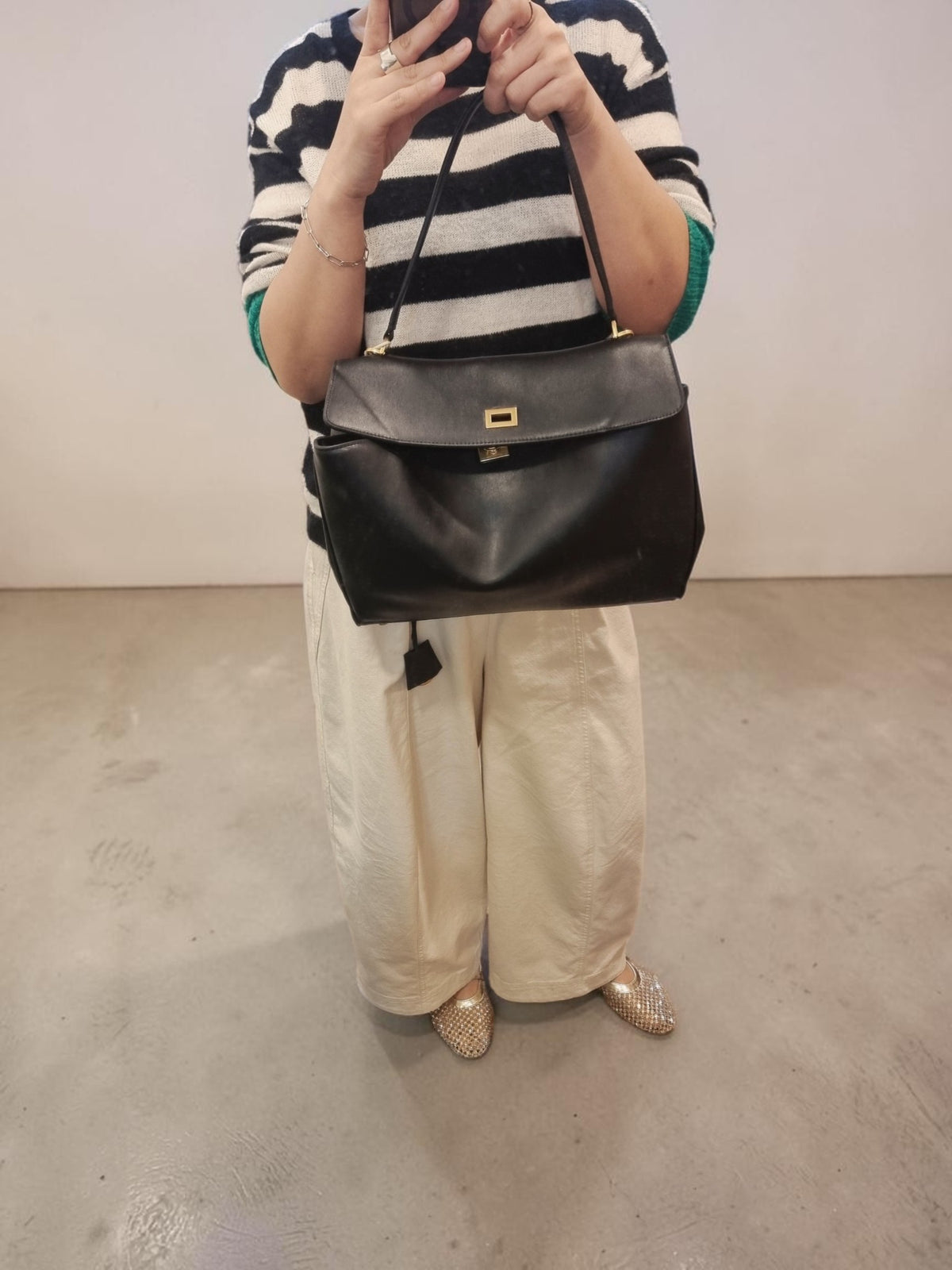 A person wearing a black and white striped sweater and cream wide-leg pants holds up a black leather shoulder bag with gold hardware against a plain indoor wall.