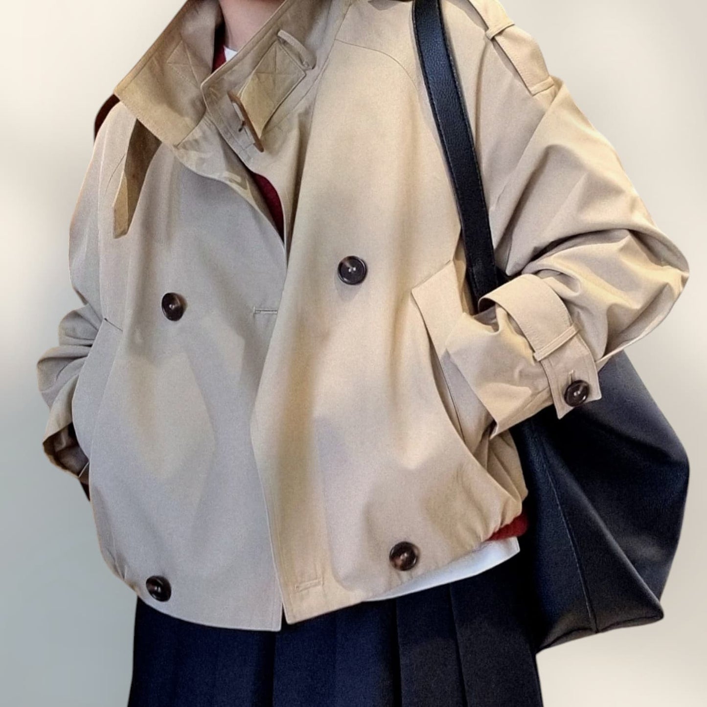 “Beige cropped trench coat with dark buttons, styled with a navy pleated skirt and black shoulder bag on a neutral background.”