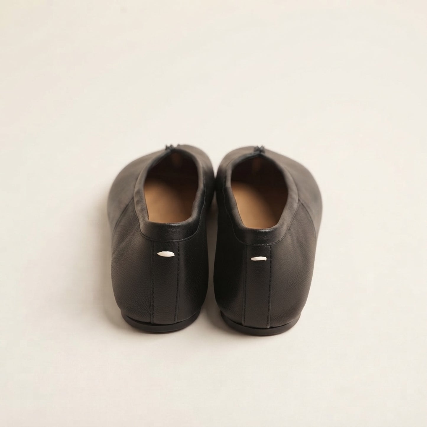 A pair of black leather ballet flats shown from the back, featuring a small silver metal accent on the heel and a tan insole, set against a plain off-white background.