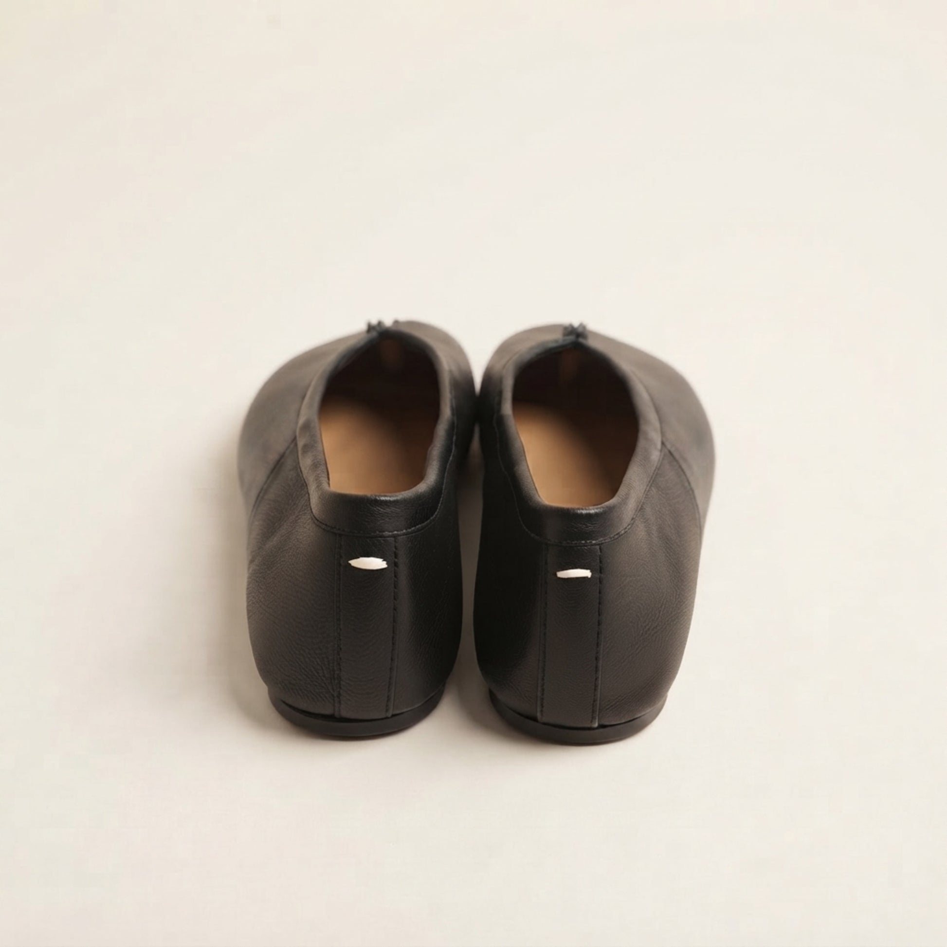 A pair of black leather ballet flats shown from the back, featuring a small silver metal accent on the heel and a tan insole, set against a plain off-white background.