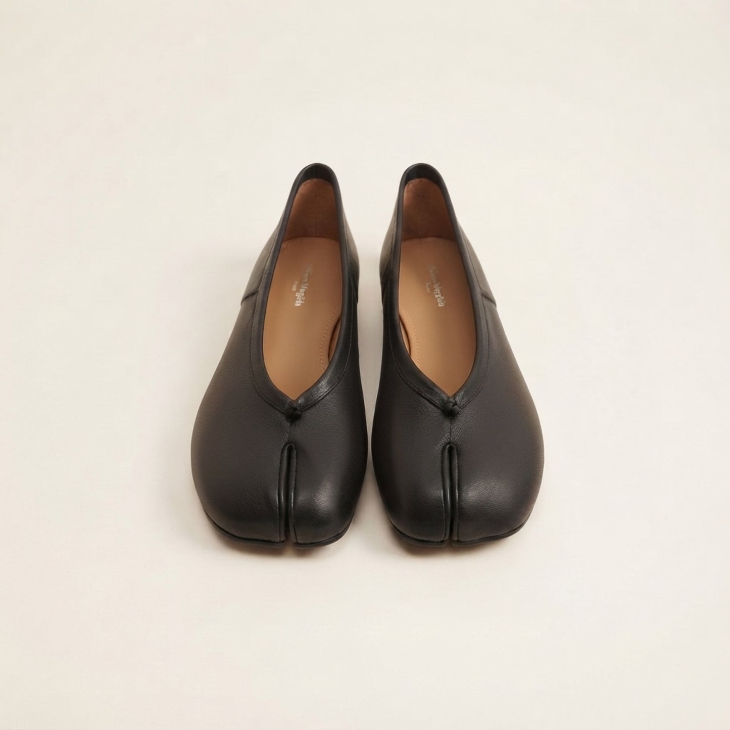 A pair of black leather Japanese tabi-style shoes with a split toe, shown from the front on a light beige background.