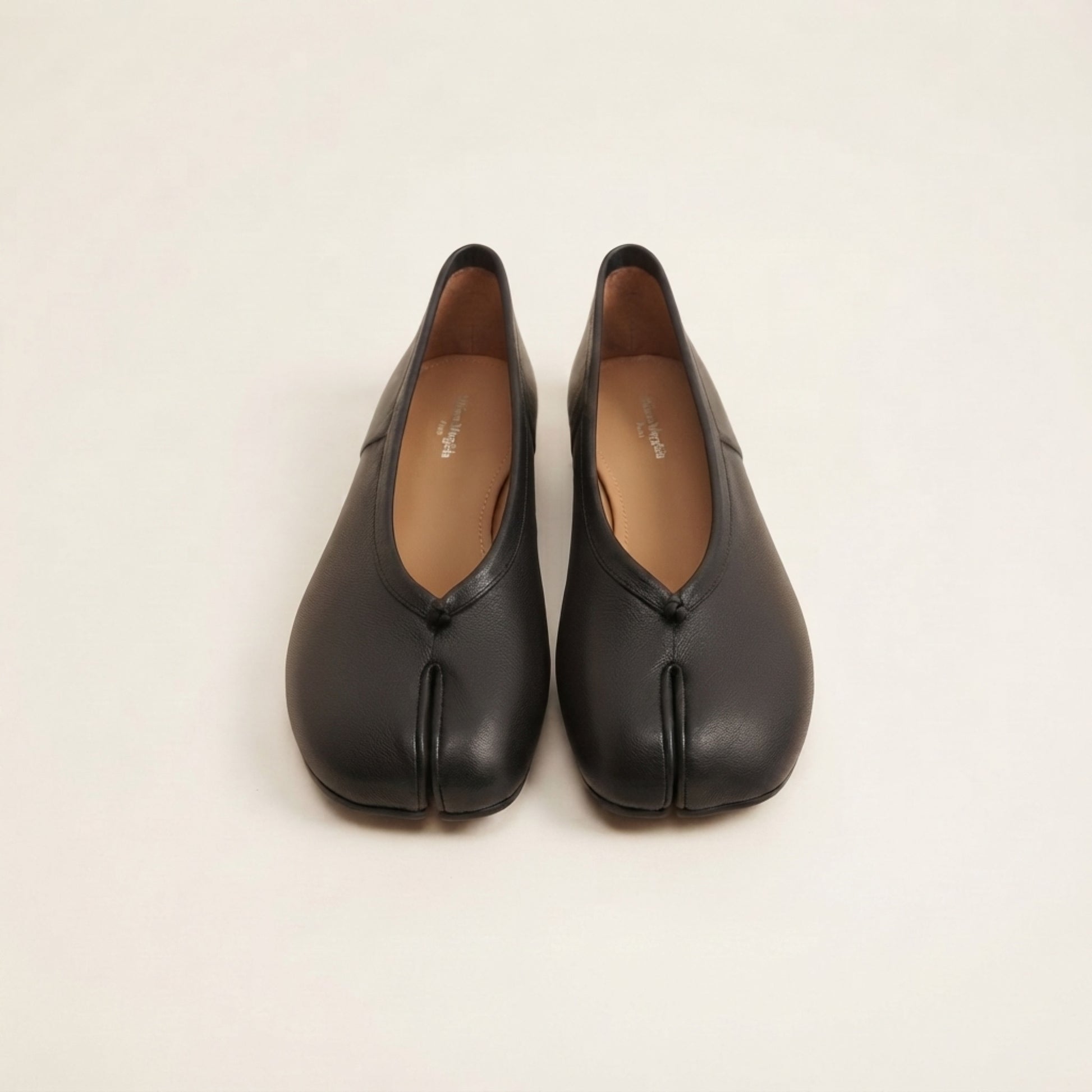 A pair of black leather Japanese tabi-style shoes with a split toe, shown from the front on a light beige background.
