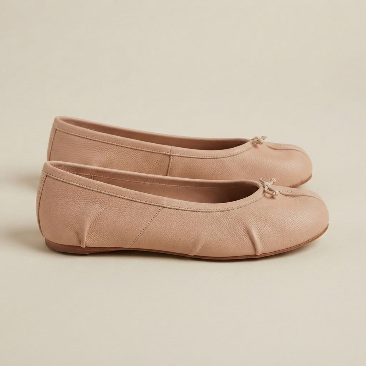 A pair of light beige leather ballet flats with a small bow detail on the toe, displayed against a neutral cream background.