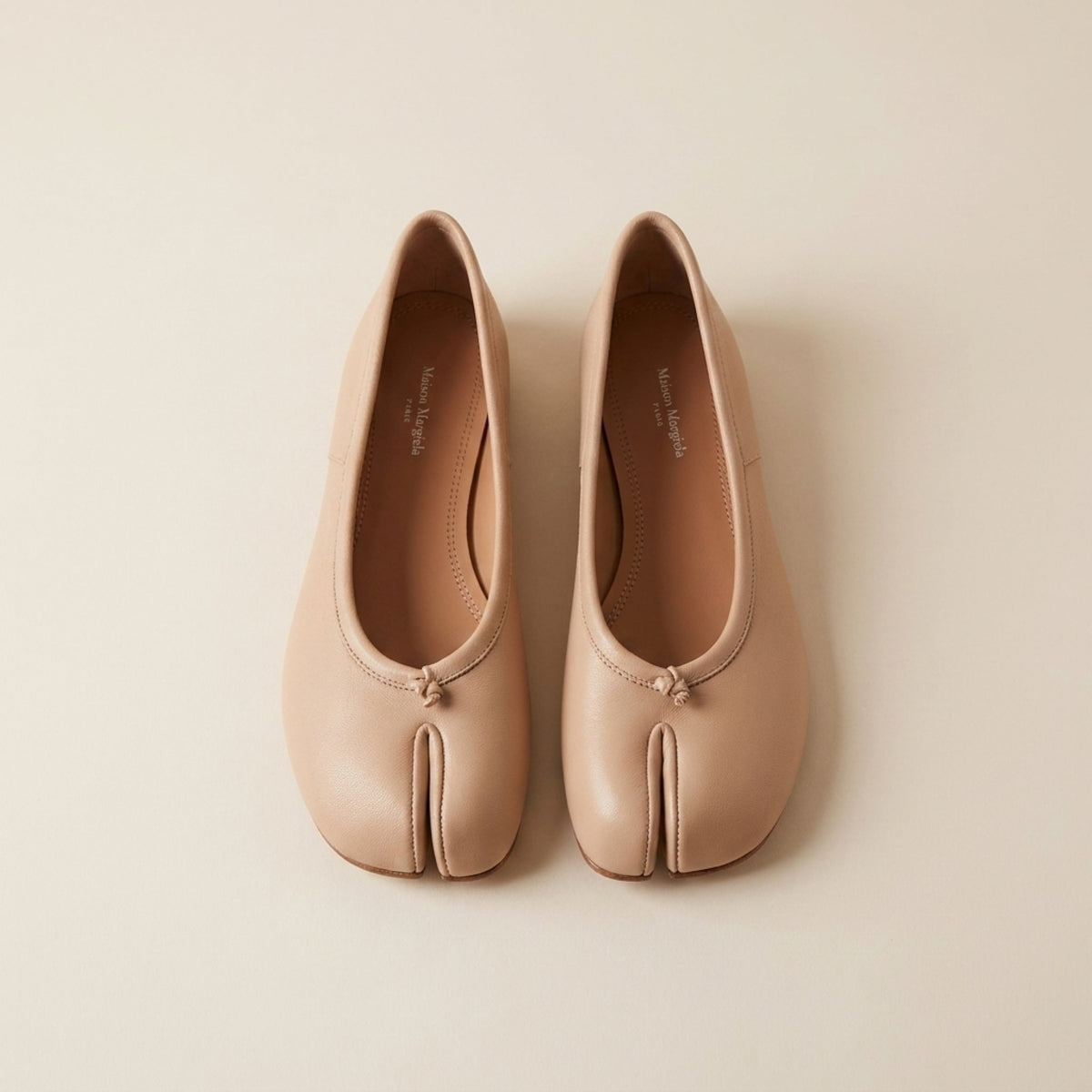 A pair of light beige leather tabi-style flats with a split toe and small front knot detail, photographed from above on a warm off-white background.